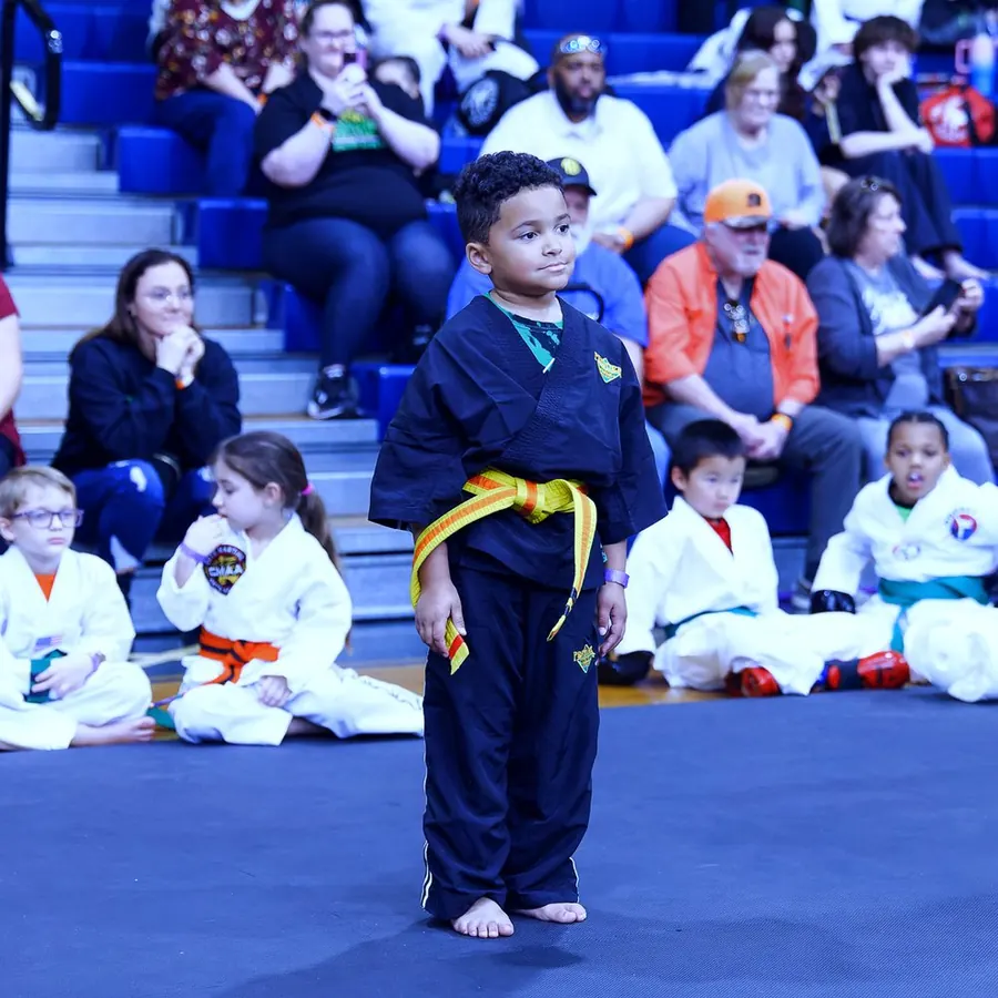 A young competitor in a black uniform and yellow-orange belt stands on the mat while other students watch from the stands.